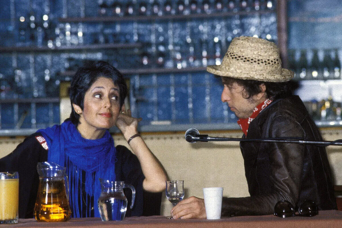 Joan Baez and Bob Dylan sit behind a table with various beverages and a microphone. She lifts her hand to her ear.