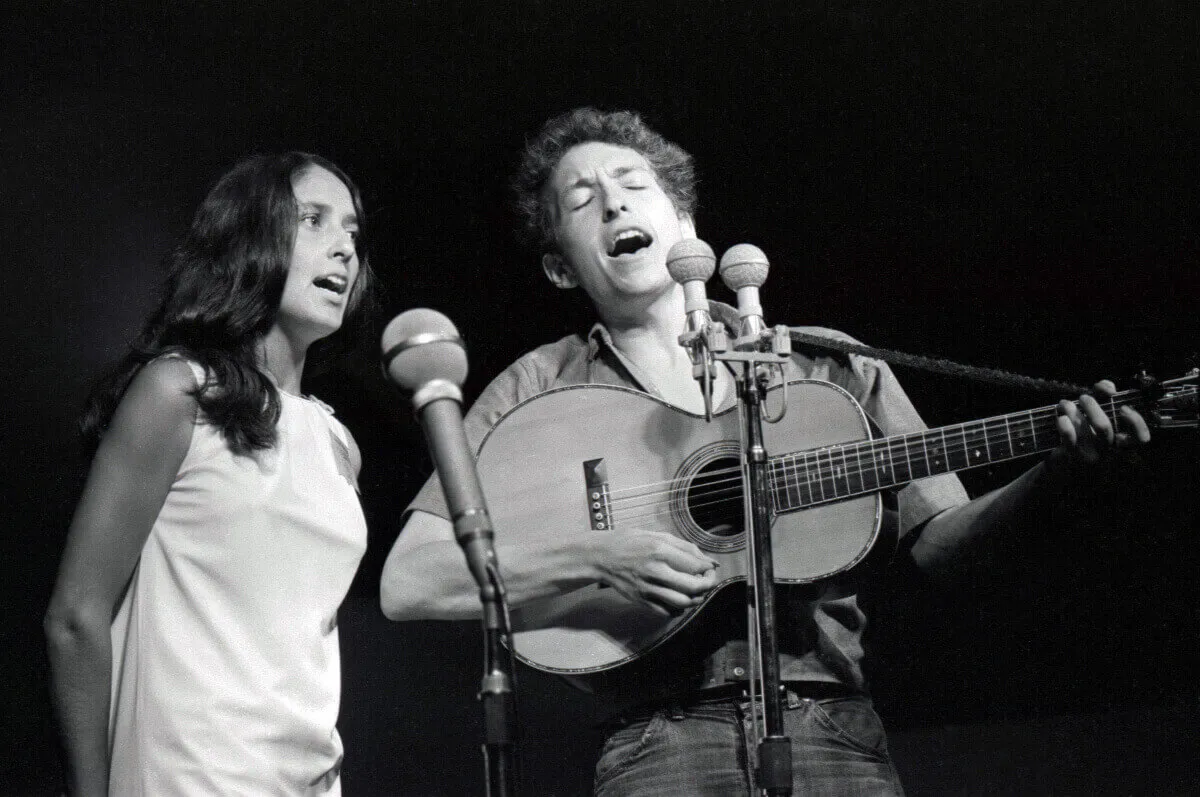 A black and white picture of Joan Baez and Bob Dylan singing into microphones. He plays guitar.