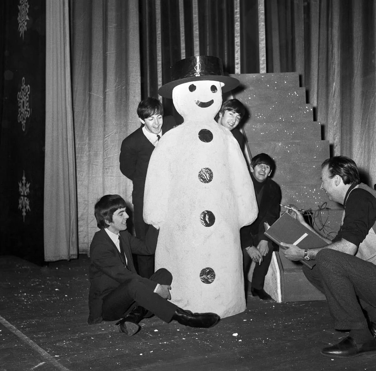 The Beatles poke out from behind a snowman statue while someone crouches near them.