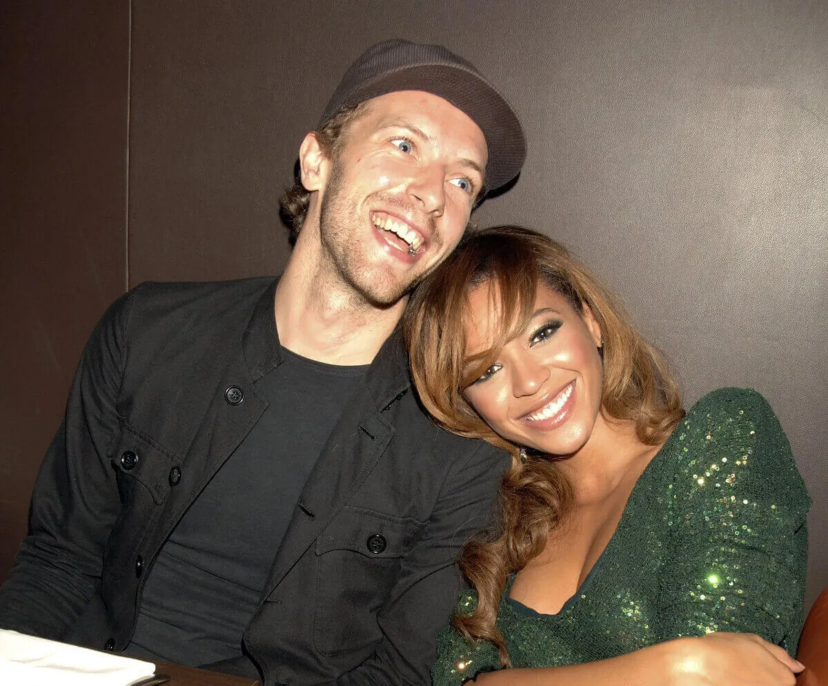 Chris Martin wears a black jacket and hat and sits with Beyonce, who wears a green shirt. She leans on his shoulder.