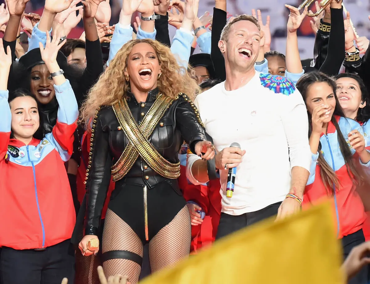 Beyonce wears a black body suit and stands with Chris Martin, who wears white. They stand with performers during the Super Bowl half time show.