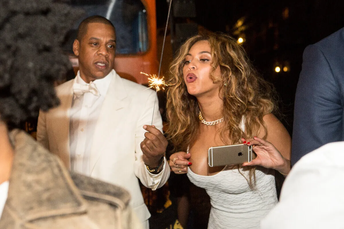 Jay-Z holds a lit sparkler and Beyonce blows on it. They both wear white.