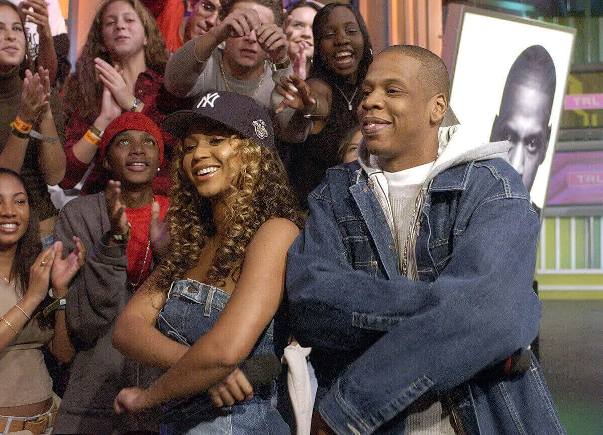 Beyonce and Jay-Z wears denim and stand with their arms folded. She wears a baseball hat.