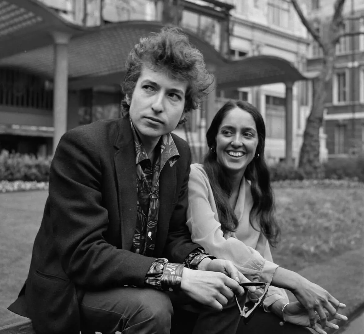 A black and white picture of Bob Dylan and Joan Baez sitting together in front of a building. She smiles.