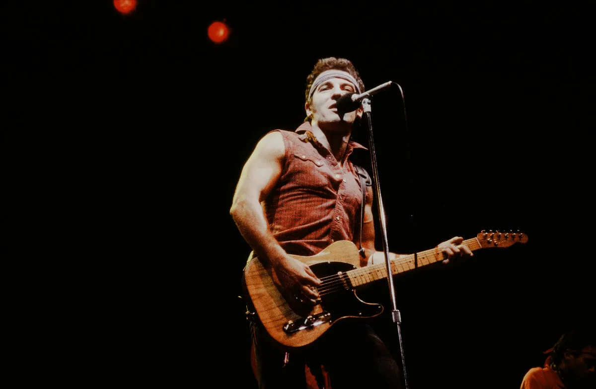 Bruce Springsteen wears a red tank top and plays guitar. He stands in front of a microphone.