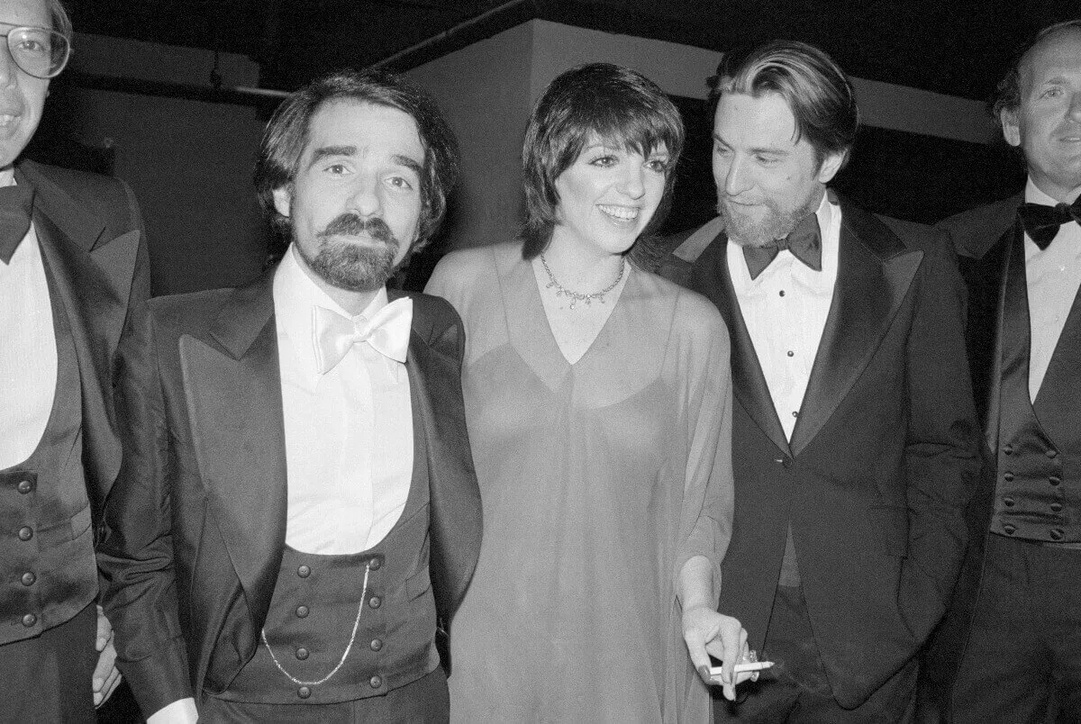 A black and white picture of Martin Scorsese, Liza Minnelli, and Robert De Niro standing together. Minnelli holds a cigarette.