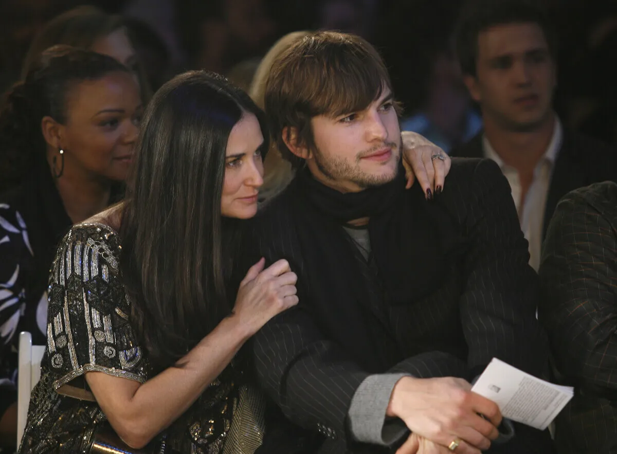 Demi Moore sits with her arm around Ashton Kutcher's shoulders. He wears a scarf.