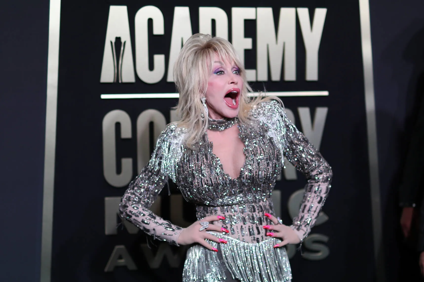 Dolly Parton with her hands on her hips looking surprised at the 2023 CMAs. She's wearing a low-cut silver fringe outfit.