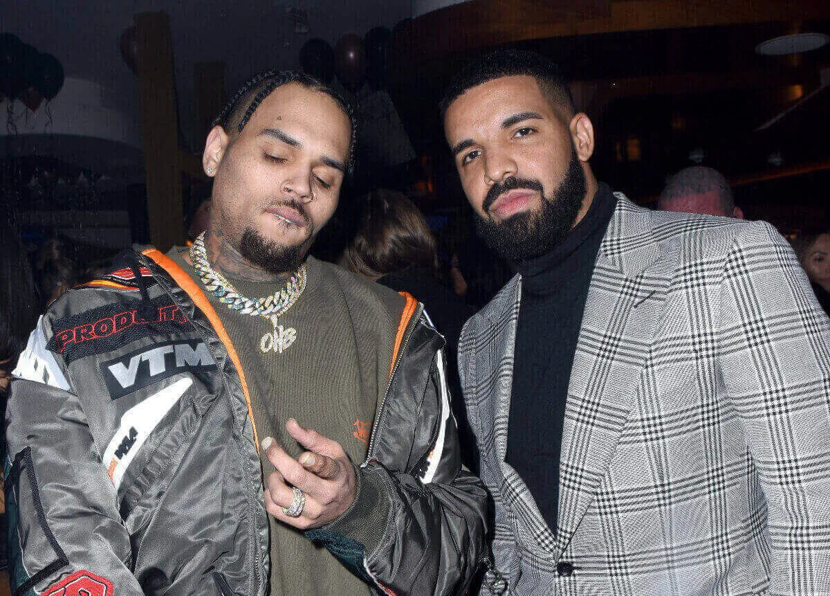 Chris Brown and Drake stand together. Brown wears a hoodie and Drake wears a plaid suit jacket.