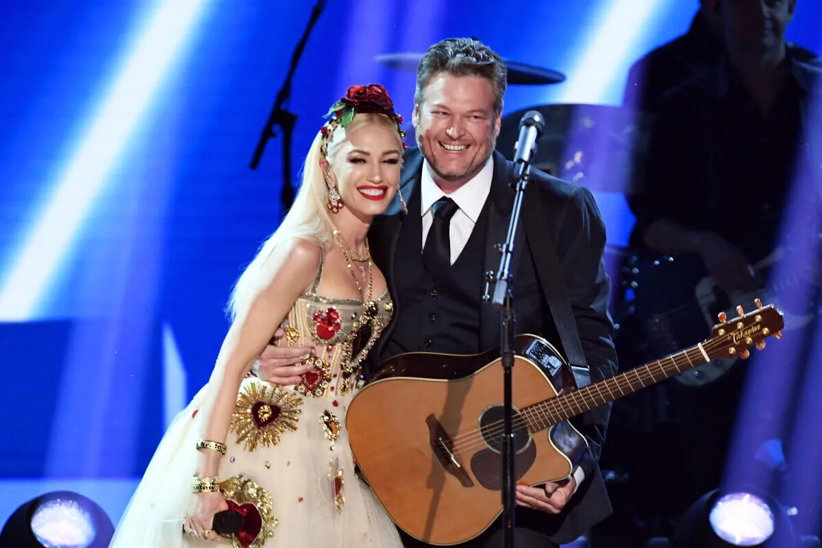 Gwen Stefani stands on a stage with Blake Shelton. She wears a dress embellished with hearts and a headpiece. Blake Shelton wears a suit and has a guitar.