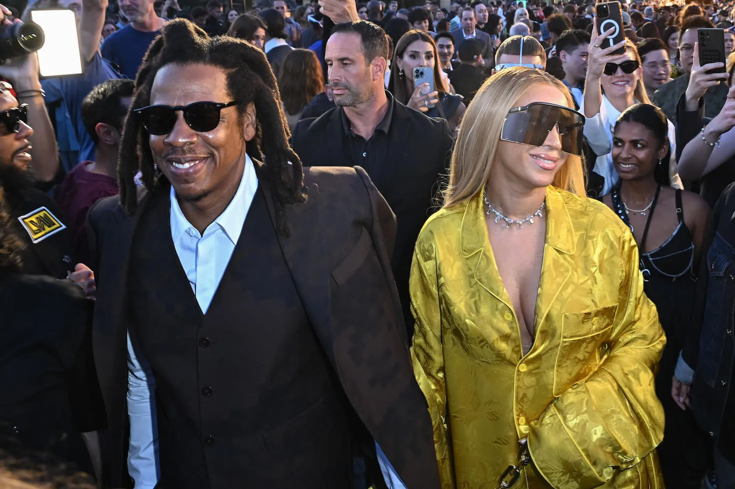 Jay-Z and Beyoncé walking in a crowd at Paris Fashion Week. Beyoncé is wearing a yellow dress with large sunglasses and Jay-Z is wearing a black suit.