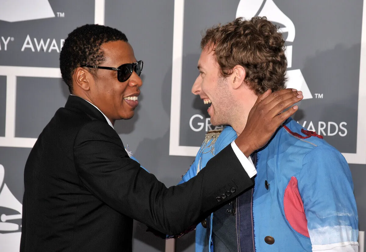 Jay-Z wears a suit and sunglasses and touches Chris Martin's shoulder. Martin wears a blue coat.