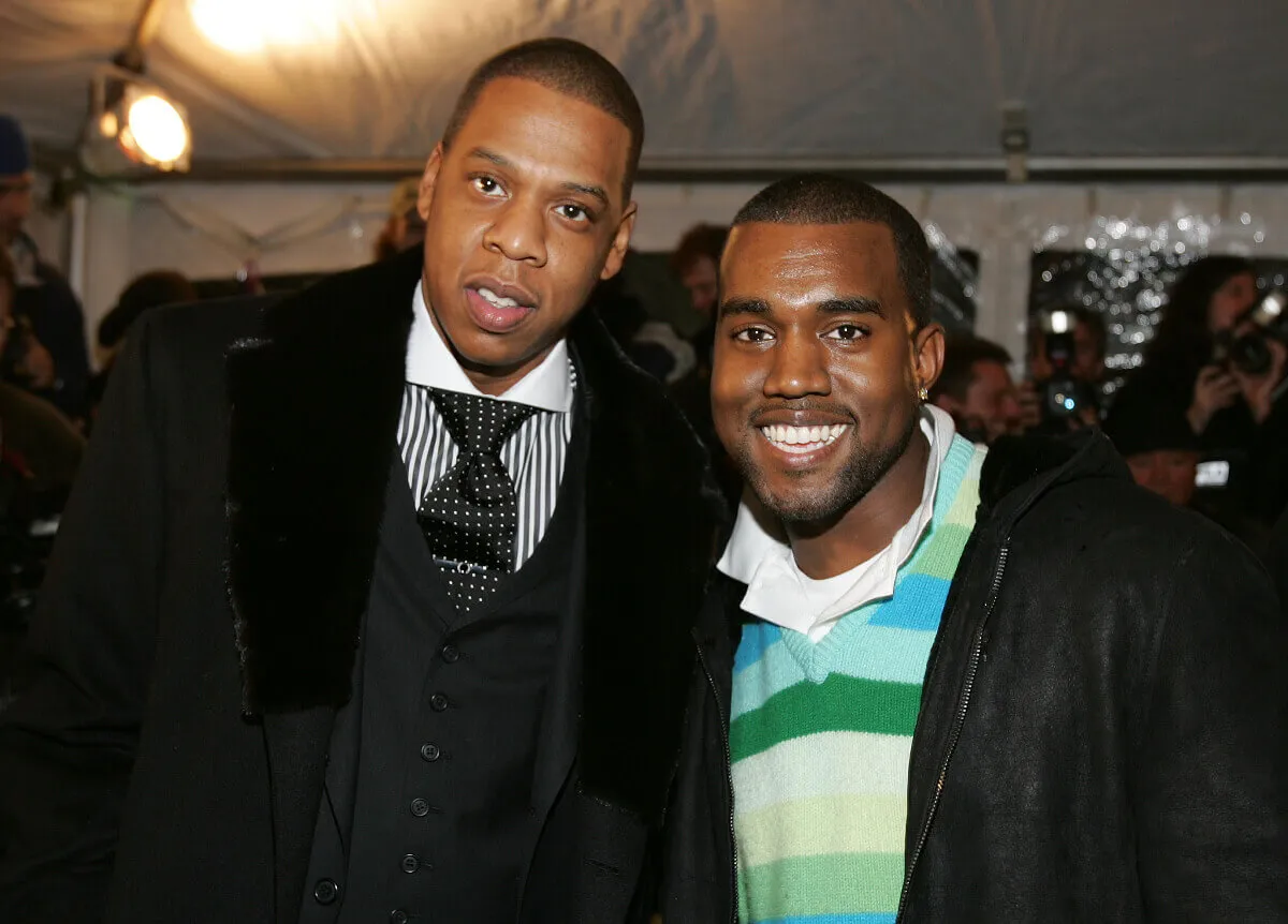 Jay Z wears a suit and stands with Kanye West, who wears a striped rugby shirt.