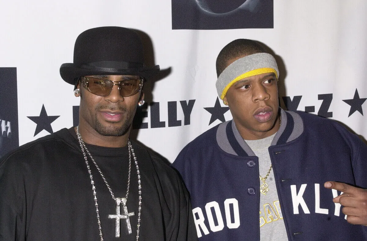 R. Kelly wears a bowler hat and sunglasses and stands with Jay-Z who wears a sweatband and a Brooklyn jacket.