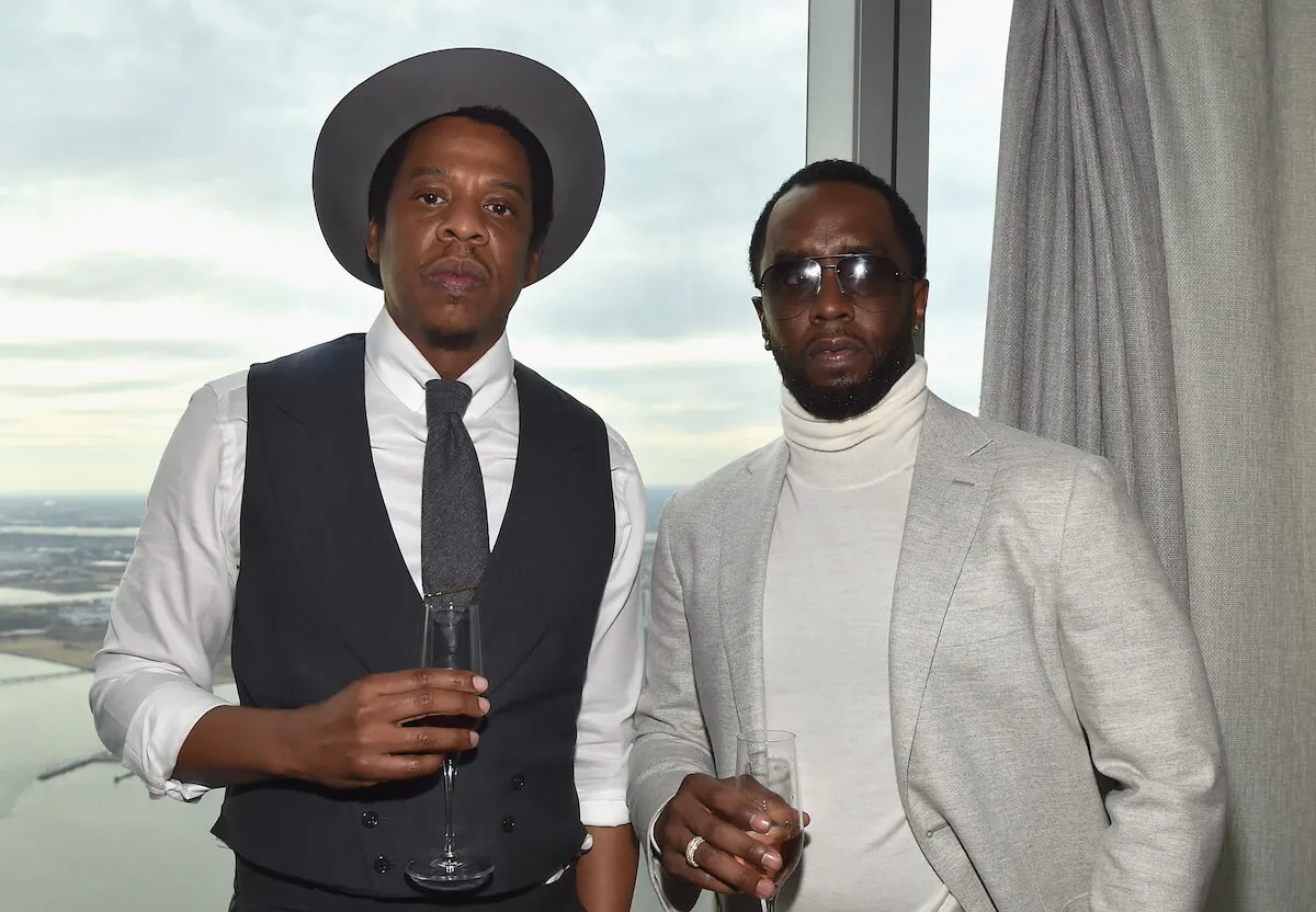 Jay-Z and Sean 'Diddy' Combs stand together and hold champagne flutes. Jay-Z wears a hat and Diddy wears sunglasses. They stand in front of a window.