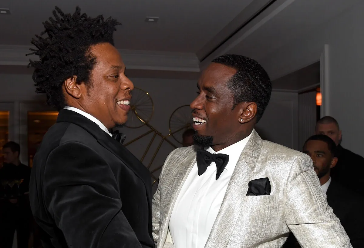 Jay-Z and Sean 'Diddy' Combs smile at each other. Jay-Z wears a black suit and Diddy wears a white tuxedo.