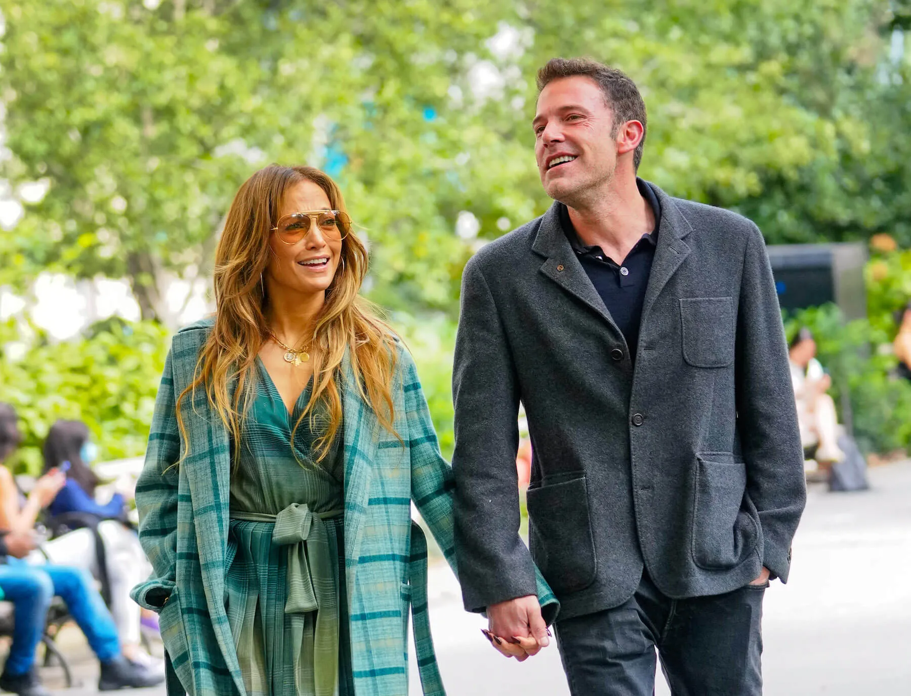 Jennifer Lopez and Ben Affleck walking hand in hand in New York City and laughing. Lopez is wearing a green plaid jacket and a green outfit underneath. Affleck is wearing a gray jacket.