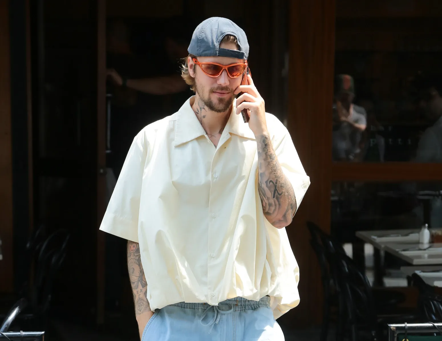 Justin Bieber holding a phone up to his ear as he walks through New York City in June 2024. He's wearing a baggy collared shirt, a backwards baseball cap, and orange sunglasses.