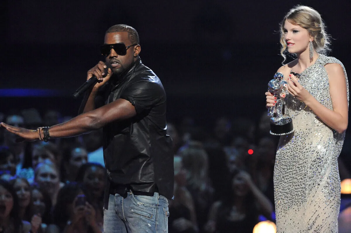 Kanye West wears a black shirt and sunglasses and talks into a microphone. Taylor Swift stands next to him in a dress, holding a VMA.