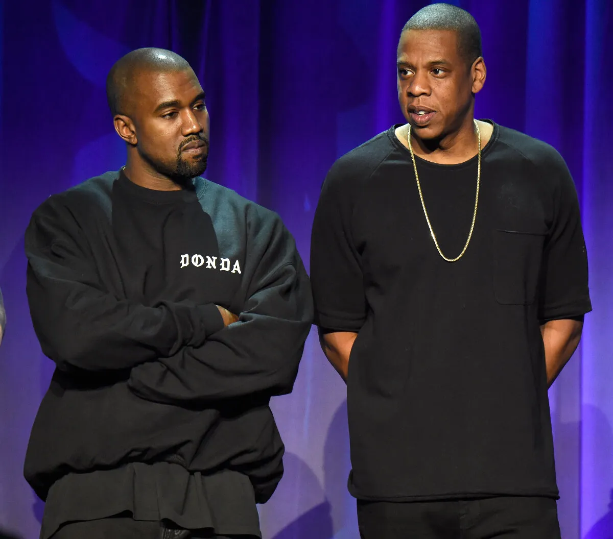 Kanye West and Jay-Z wear black shirts and stand together. West has his arms crossed.