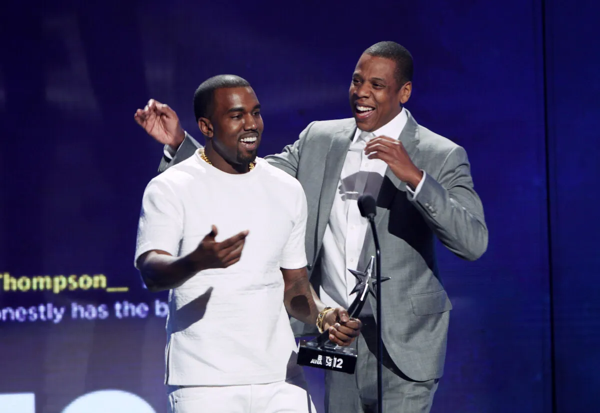 Kanye West and Jay-Z stand together. West wears a white shirt and Jay-Z wears a gray suit. They laugh.