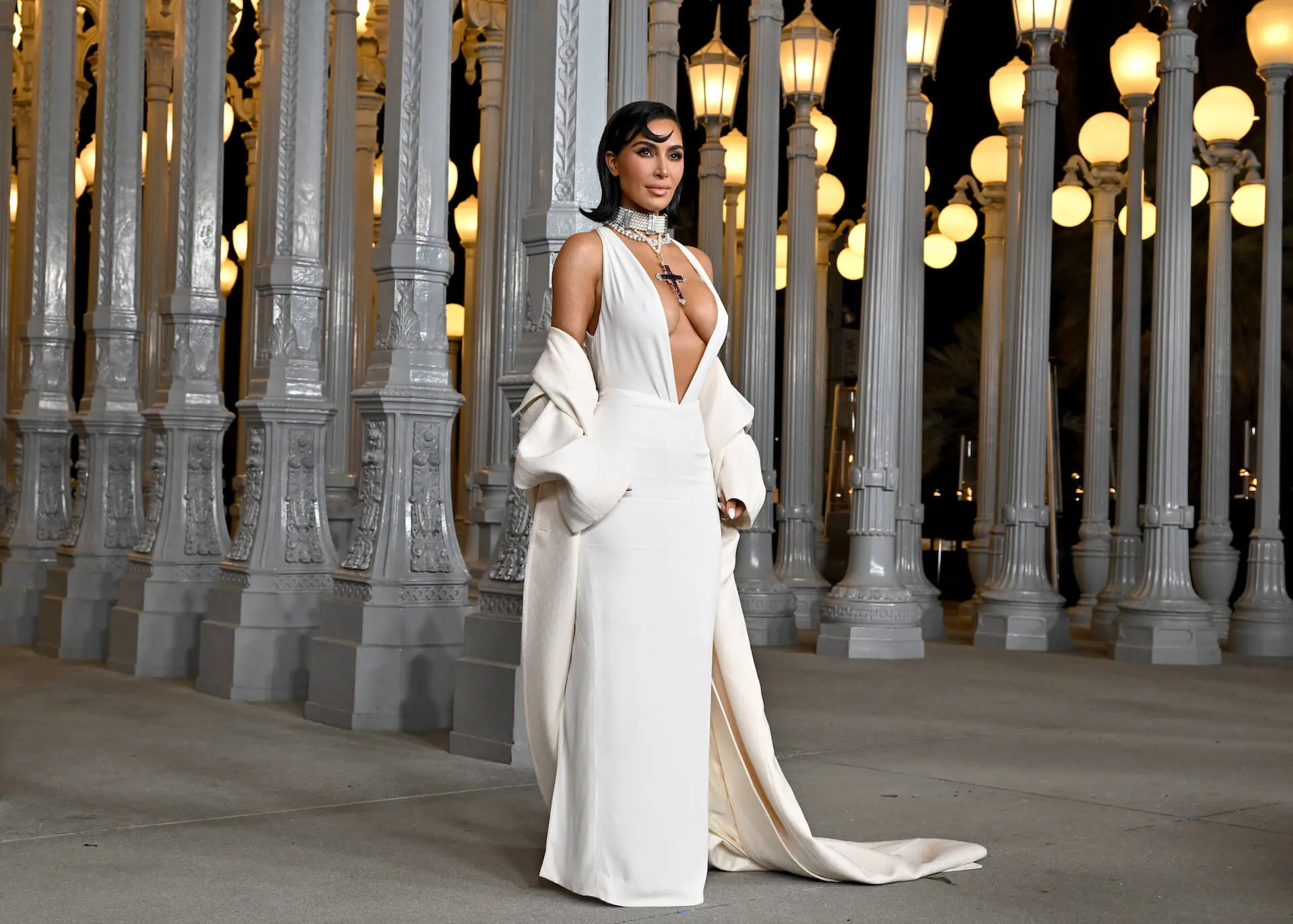 Kim Kardashian posing at the Los Angeles County Museum of Art in a white, low-cut gown with an off-the-shoulder white coat. Her hair is shoulder-length and curled up at the ends.