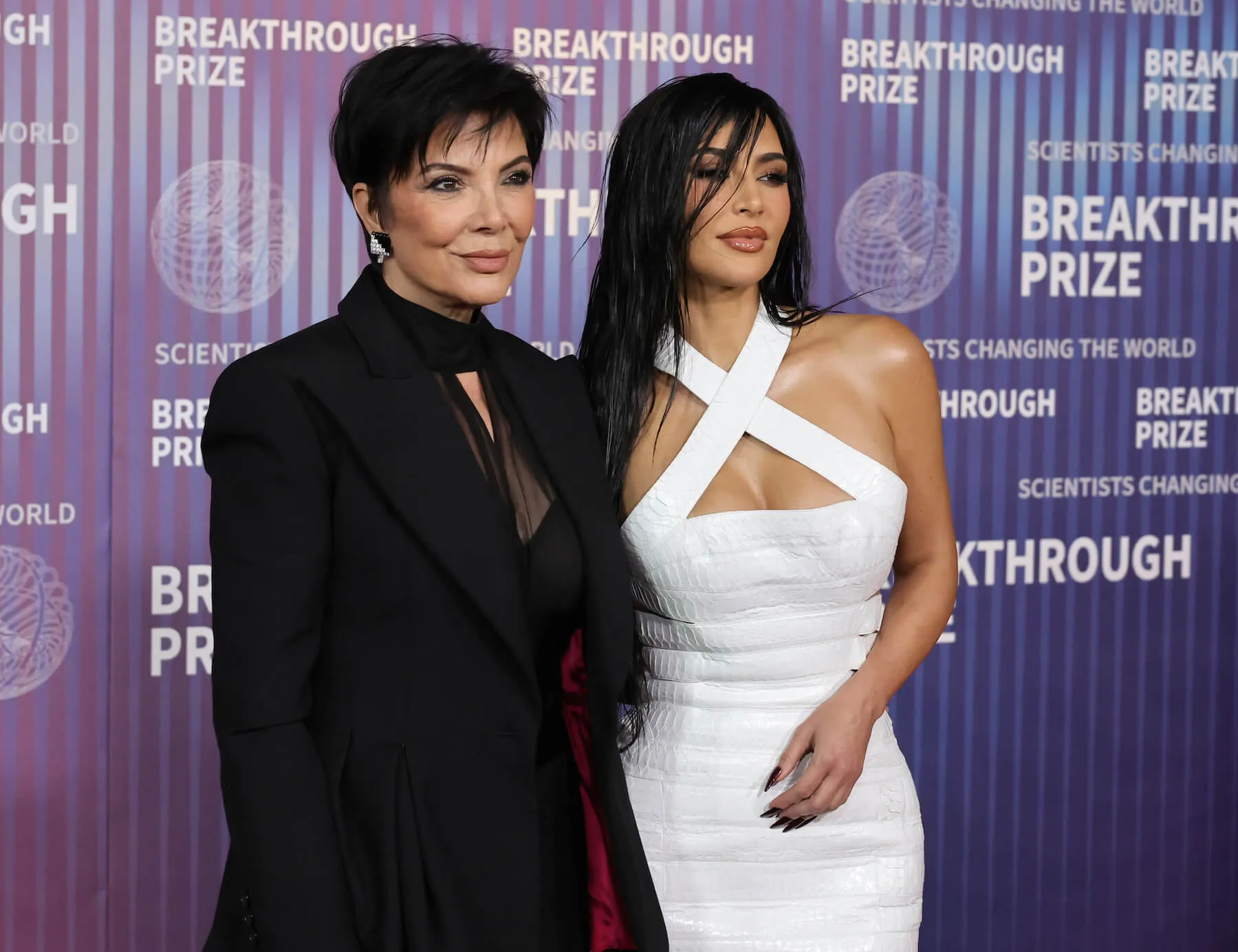 Kris Jenner in a black dress and jacket standing with Kim Kardashian in a white strapped cross front dress