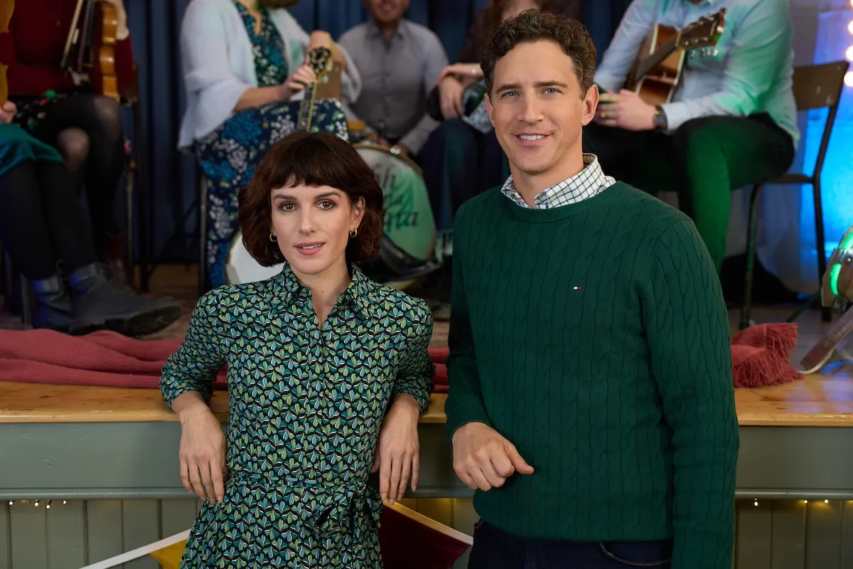 A woman with short dark hair in a green patterned dress standing next to a man in a green sweater in the Hallmark Channel movie 'Love of the Irish'