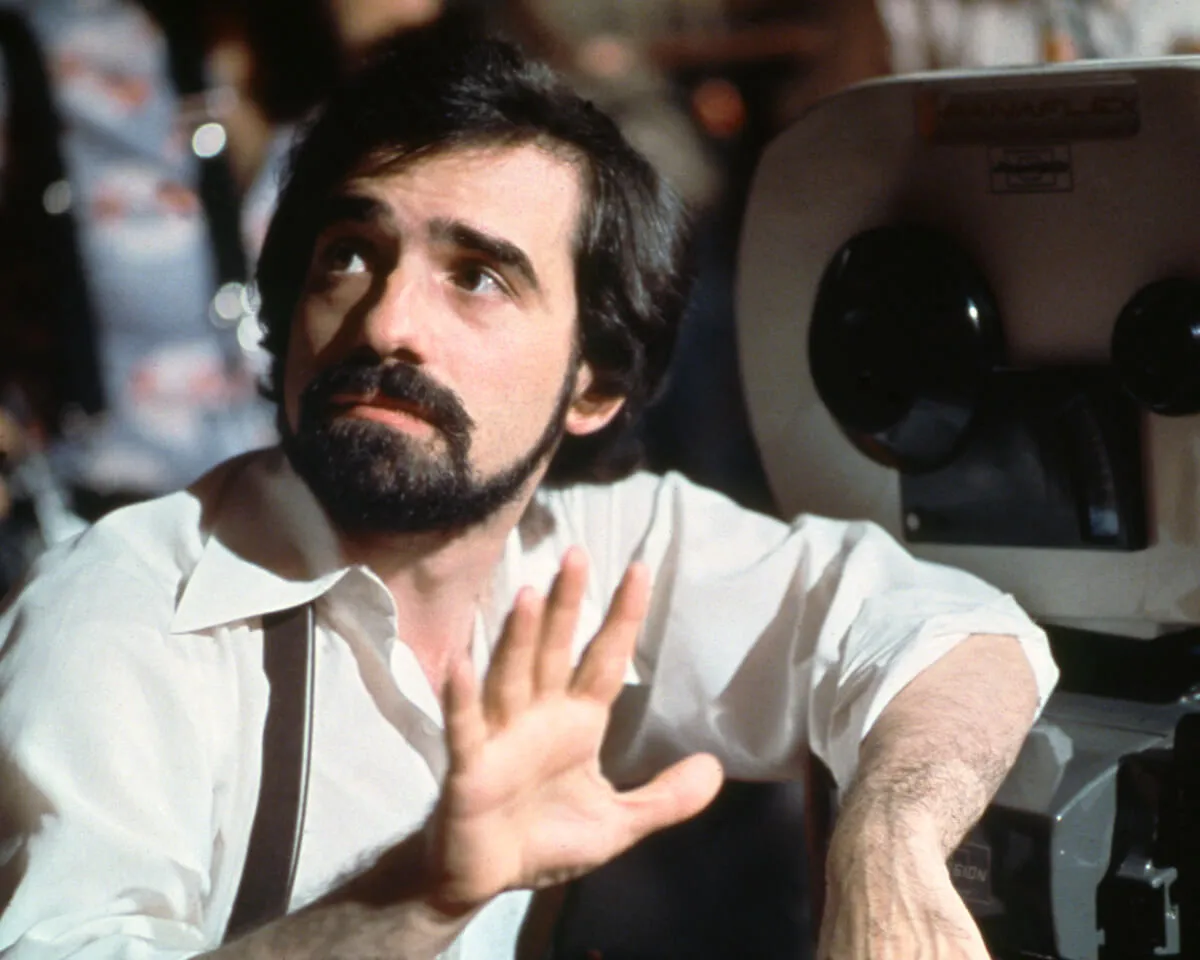 Martin Scorsese wears a white collared shirt and sits. He leans his arm on a camera.