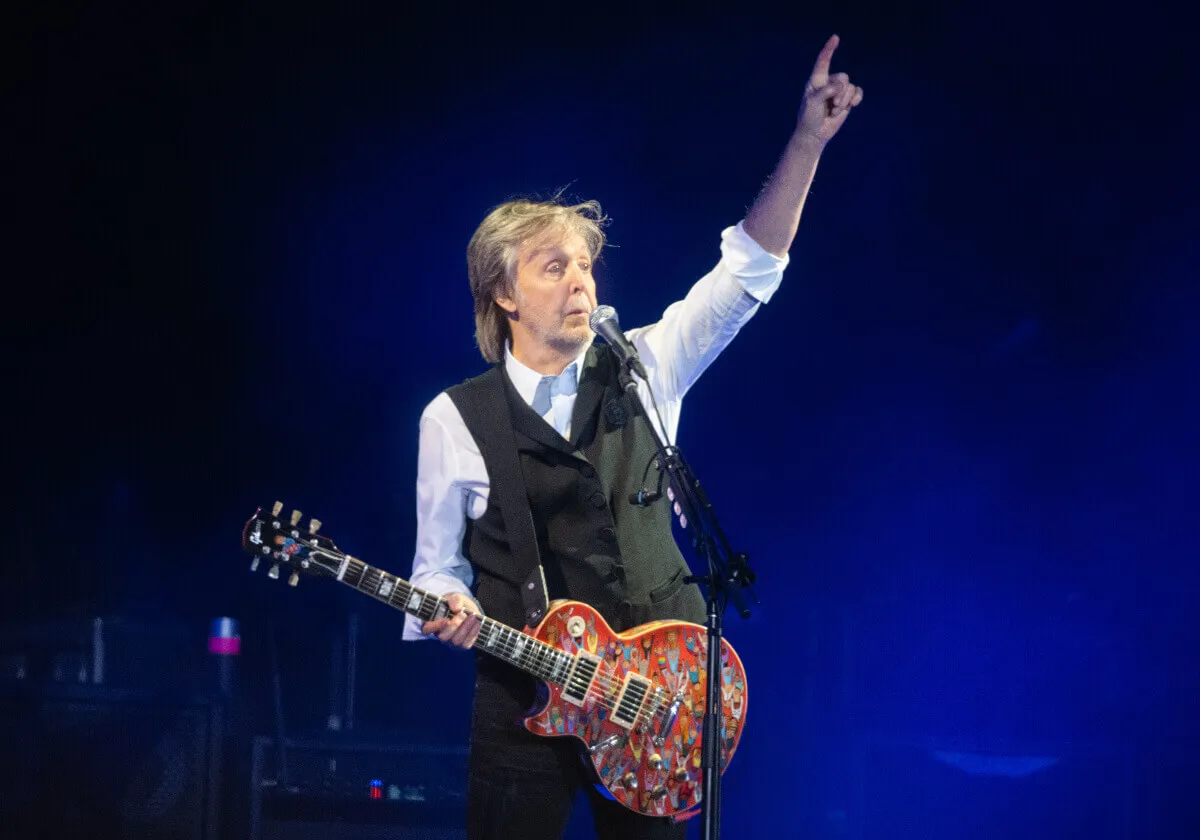 Paul McCartney holds a guitar and stands in front of a microphone with his arm up.