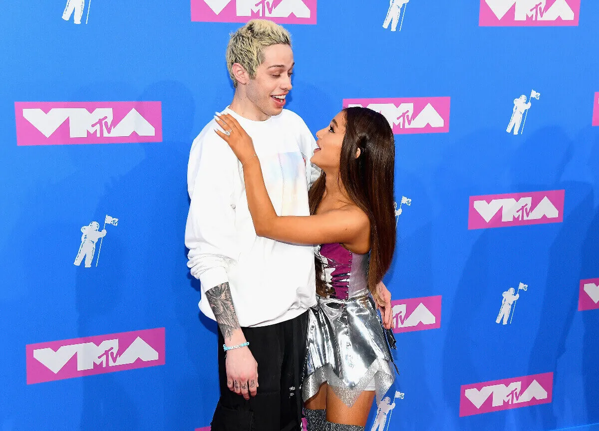 Pete Davidson and Ariana Grande Grande embrace and stand in front of a blue background. He wears a white shirt and she wears a silver dress.