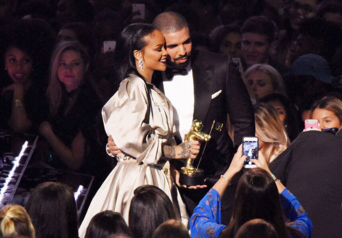 Rihanna and Drake embrace in front of a crowd. She wears a gold dress and holds a VMA and he wears a tuxedo.