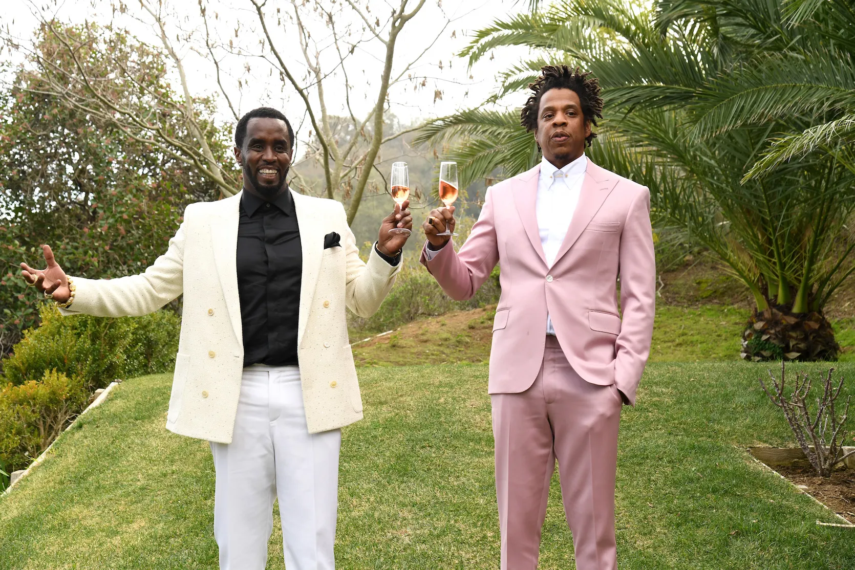 Sean 'Diddy' Combs in a yellow blazer holding a glass of champagne next to Jay-Z in a pink suit also holding up a glass of champagne. The men are outside surrounded by trees and grass.