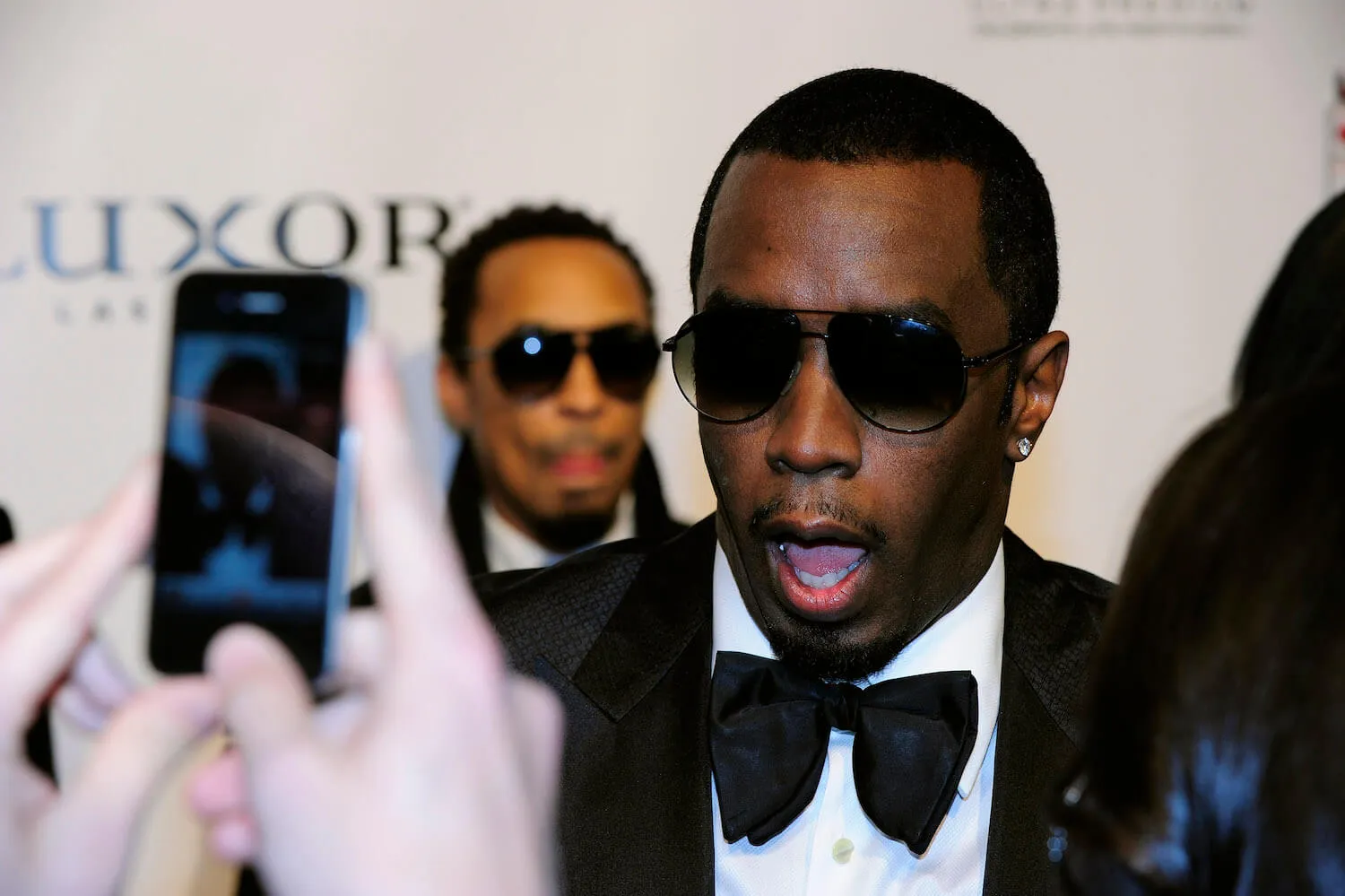 Sean 'Diddy' Combs wearing sunglasses and a black suit with a bow tie and white undershirt. His mouth is open as a phone points at him to take a photo.