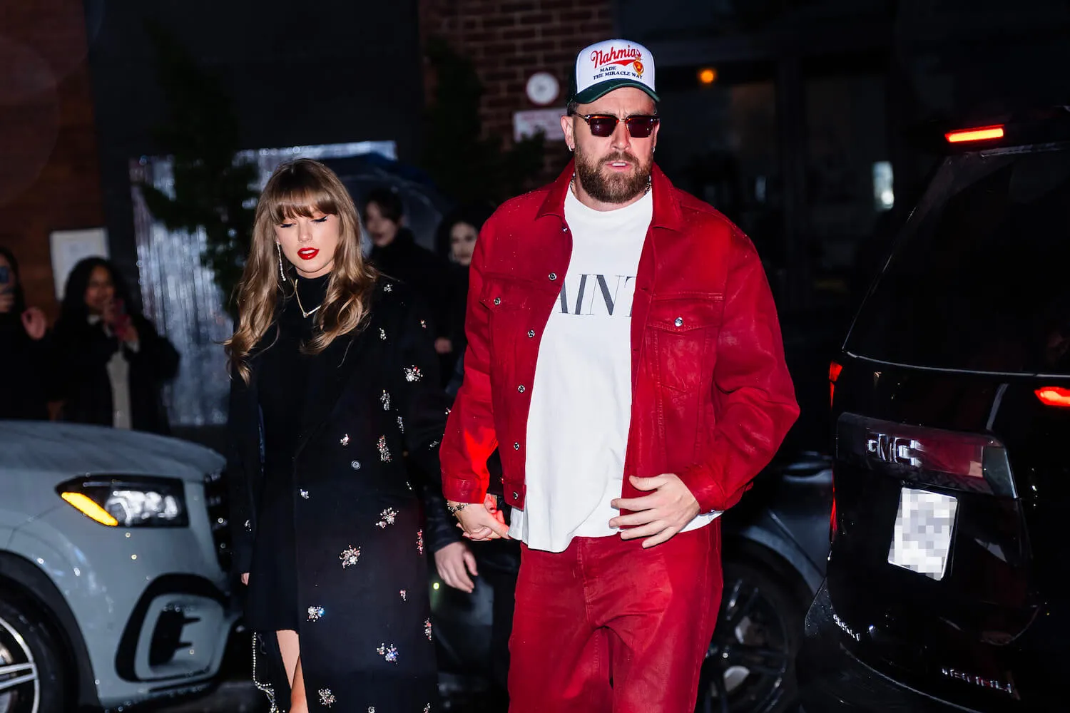 Taylor Swift in a dark dress walking with Travis Kelce at night in New York City, who's dressed in a red jacket and matching pants