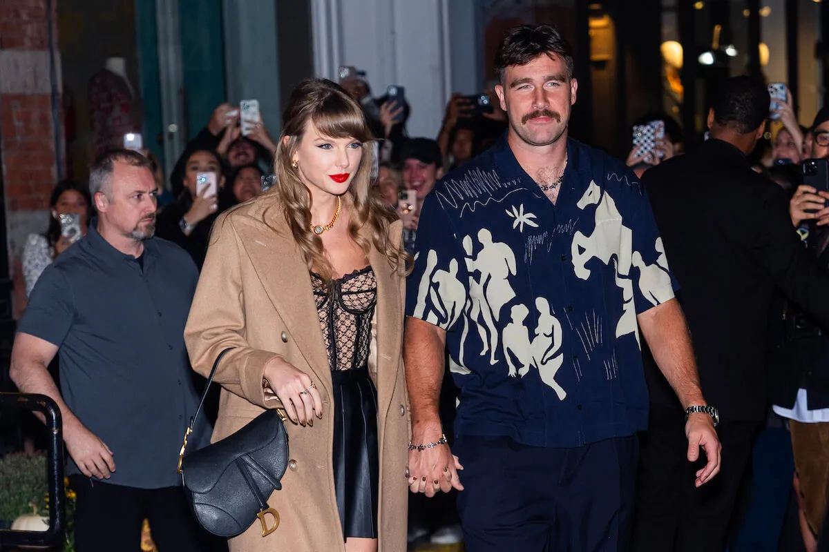 Taylor Swift and Travis Kelce holding hands while walking through a crowd. Swift is wearing a brown coat and fitted dress. Kelce is wearing a blue short-sleeve patterned shirt.