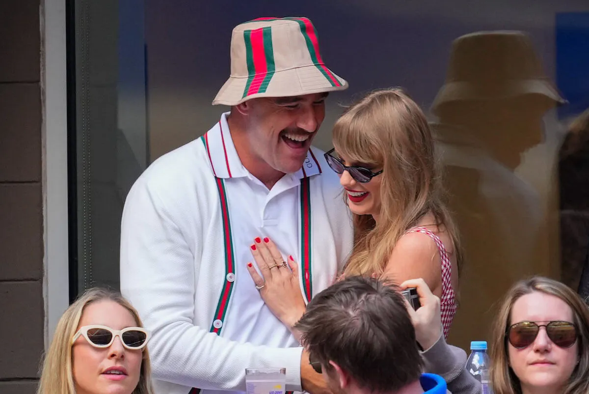 Travis Kelce and Taylor Swift leaning into each other and laughing at the US Open. Kelce is wearing a bucket hat with matching suspenders. Swift is wearing a sleeveless red plaid dress.