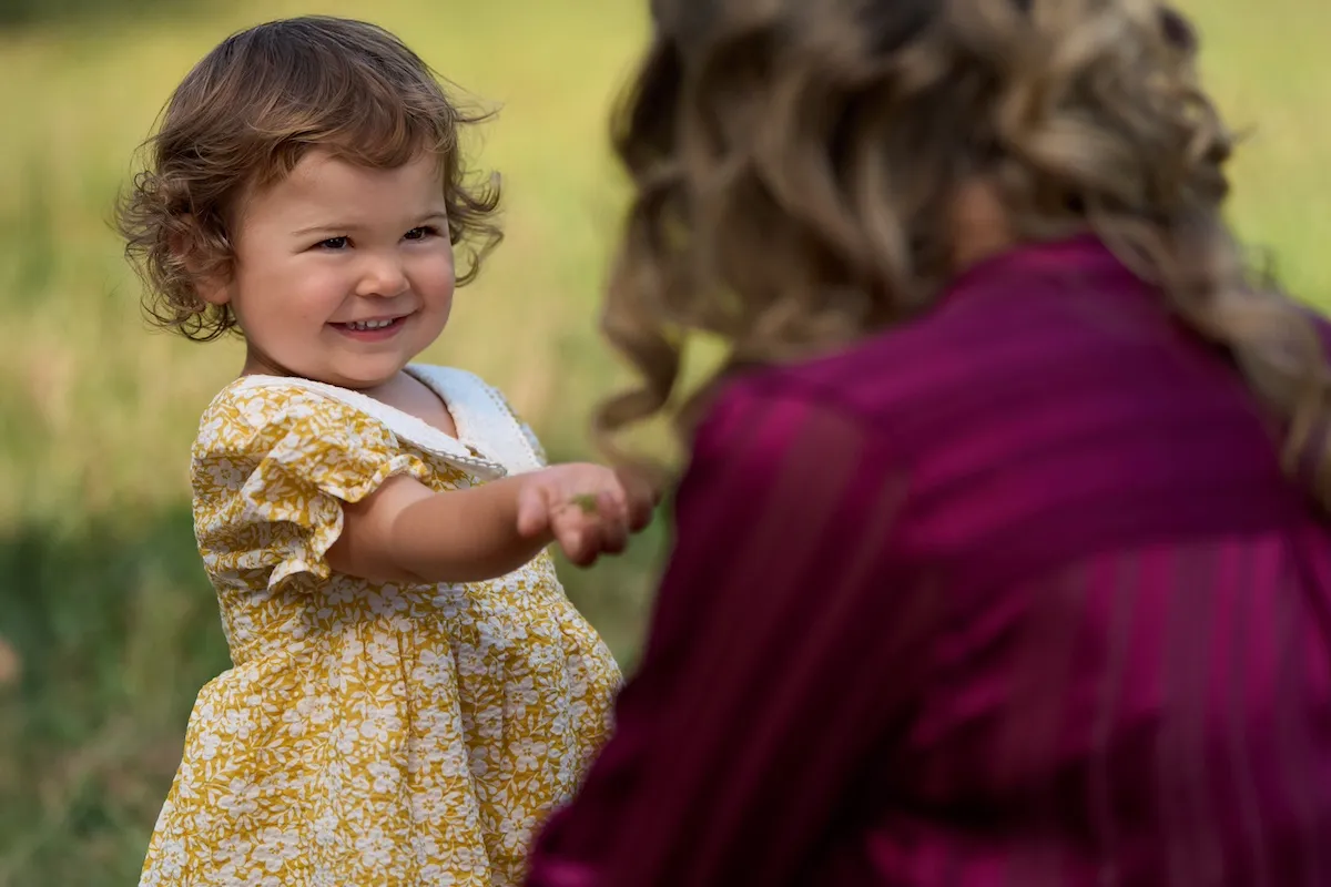 A smiling toddler girl in a yellow dress reaches out to a woman in a plum-colored dress