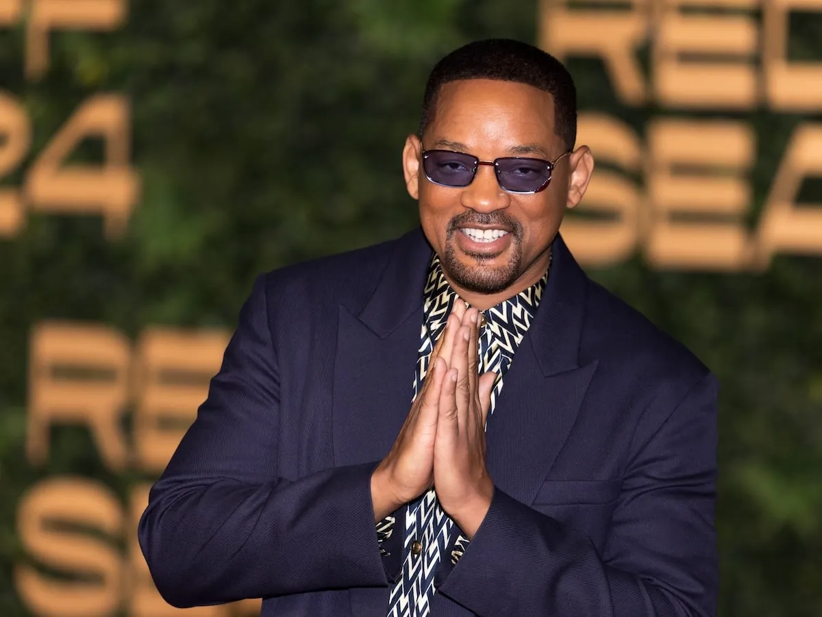 Wearing a navy suit and blue sunglasses, Will Smith puts his hands in a prayer position while taking photos on the red carpet at the Opening Ceremony of The Red Sea International Film Festival 2024