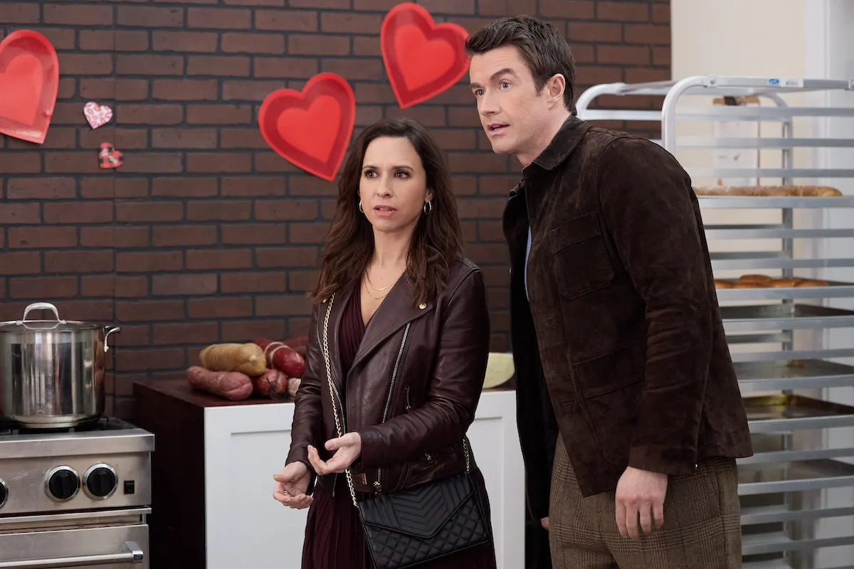 Lacey Chabert and Robert Buckley standing in front of a black brick wall decorated with red hearts in the Hallmark movie 'An Unexpected Valentine'