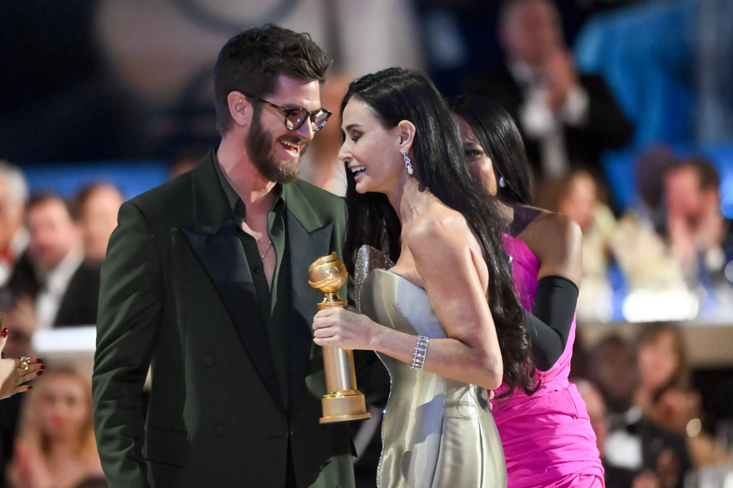 Andrew Garfield facing Demi Moore with his arm around her waist at the Golden Globes in 2025. She's holding a Golden Globe award and smiling.