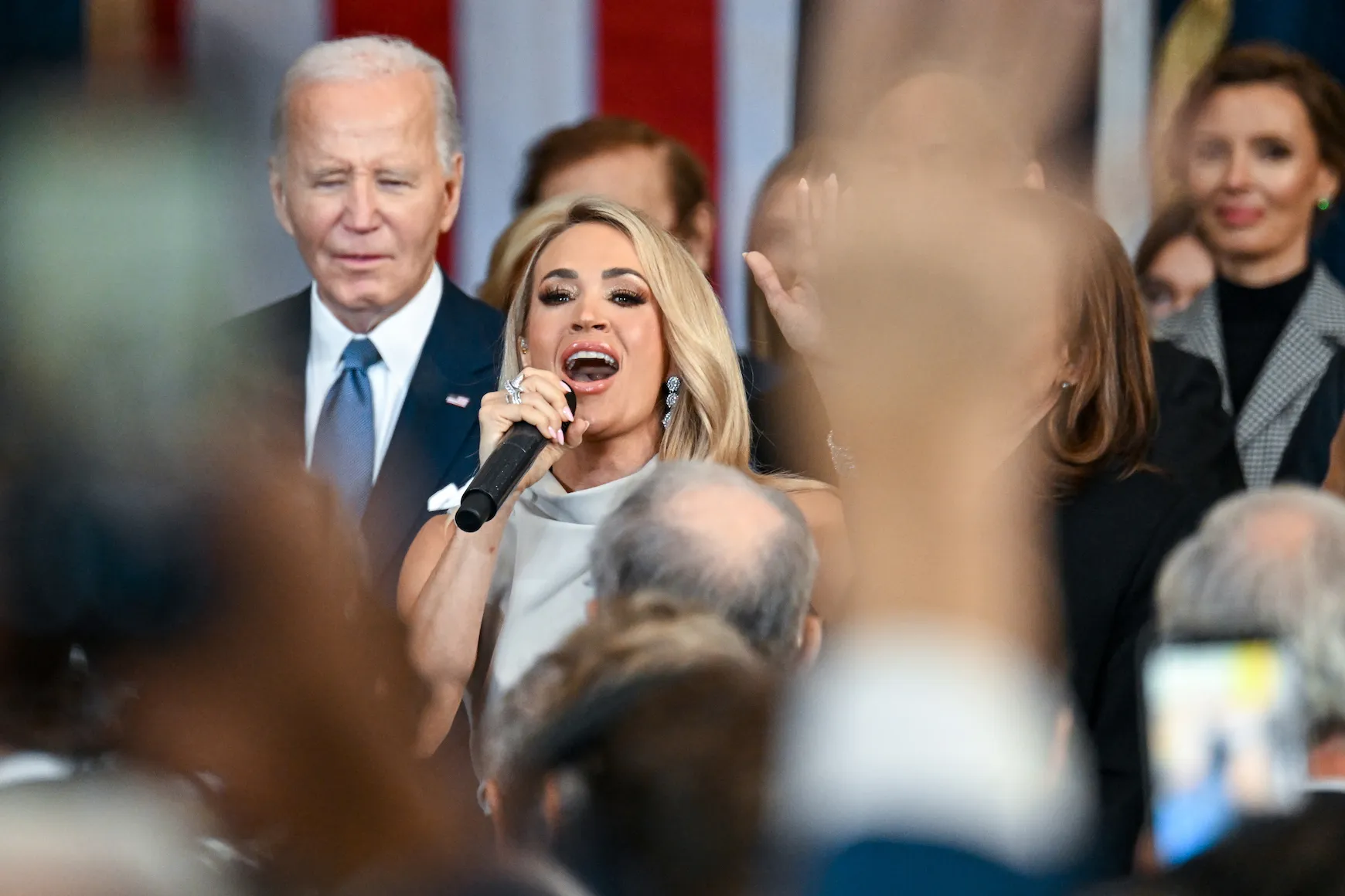 Carrie Underwood holding a microphone to her mouth and singing with her left hand up amongst a crowd of politicians. 