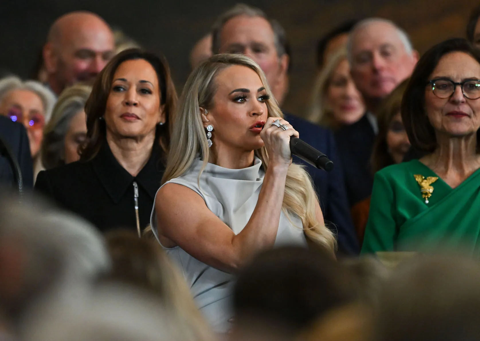 Carrie Underwood surrounded by politicians at the inauguration of Donald Trump. She's holding a microphone to her mouth as she sings.