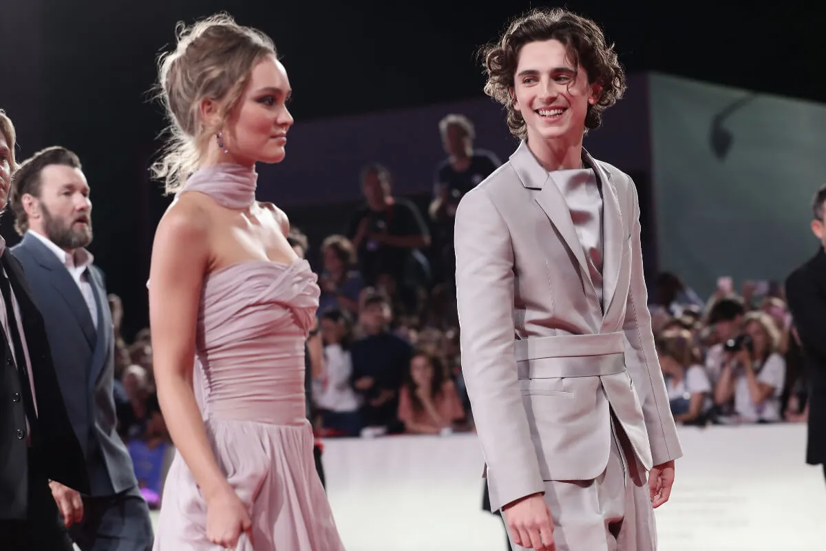 Timothée Chalamet and Lily-Rose Depp stand on a red carpet together. He wears a gray suit and she wears a pink dress.