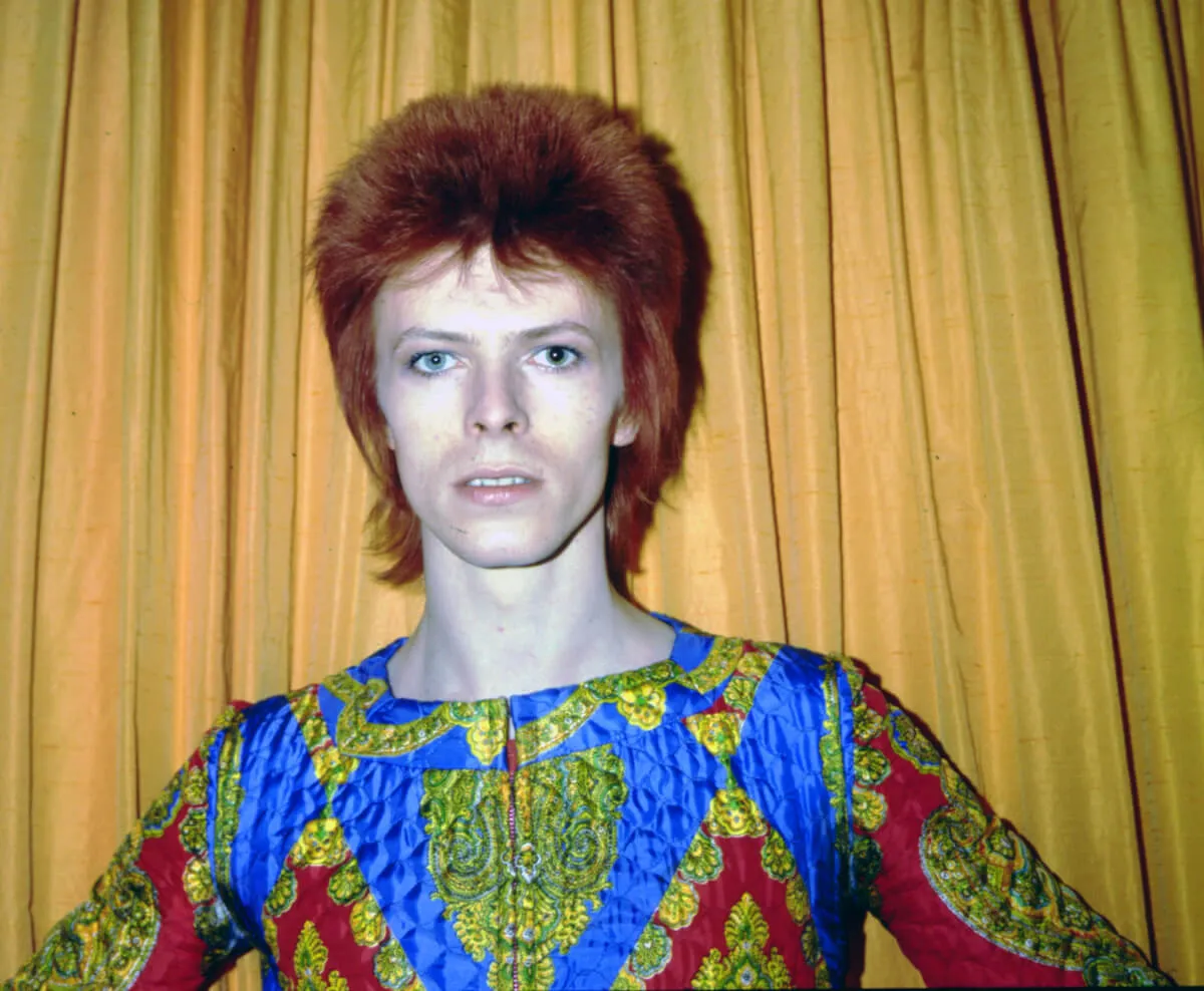 David Bowie wears a patterned shirt and stands with his hands on his hips in front of a yellow curtain.