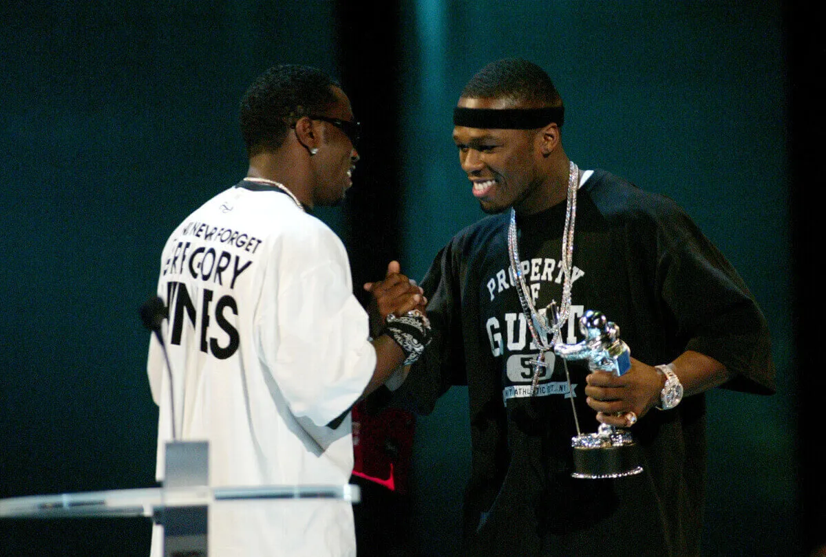 Diddy wears a white shirt and clasps 50 Cent's hand. 50 Cent wears a black shirt and holds a trophy.