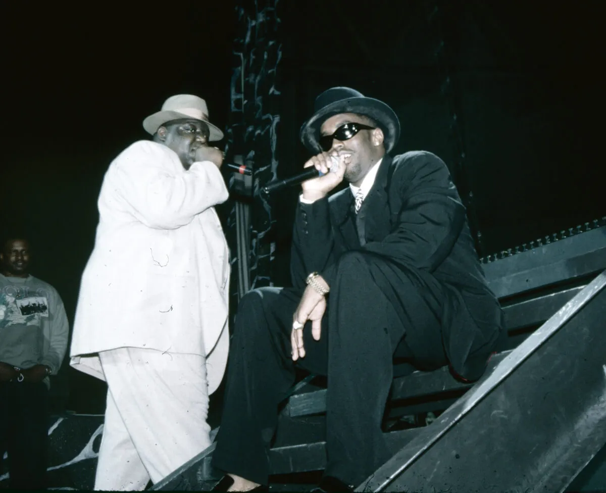 The Notorious B.I.G. wears a white suit and hat and holds a microphone. Sean 'Diddy' Combs wears a black suit and hat and sits, holding a microphone.