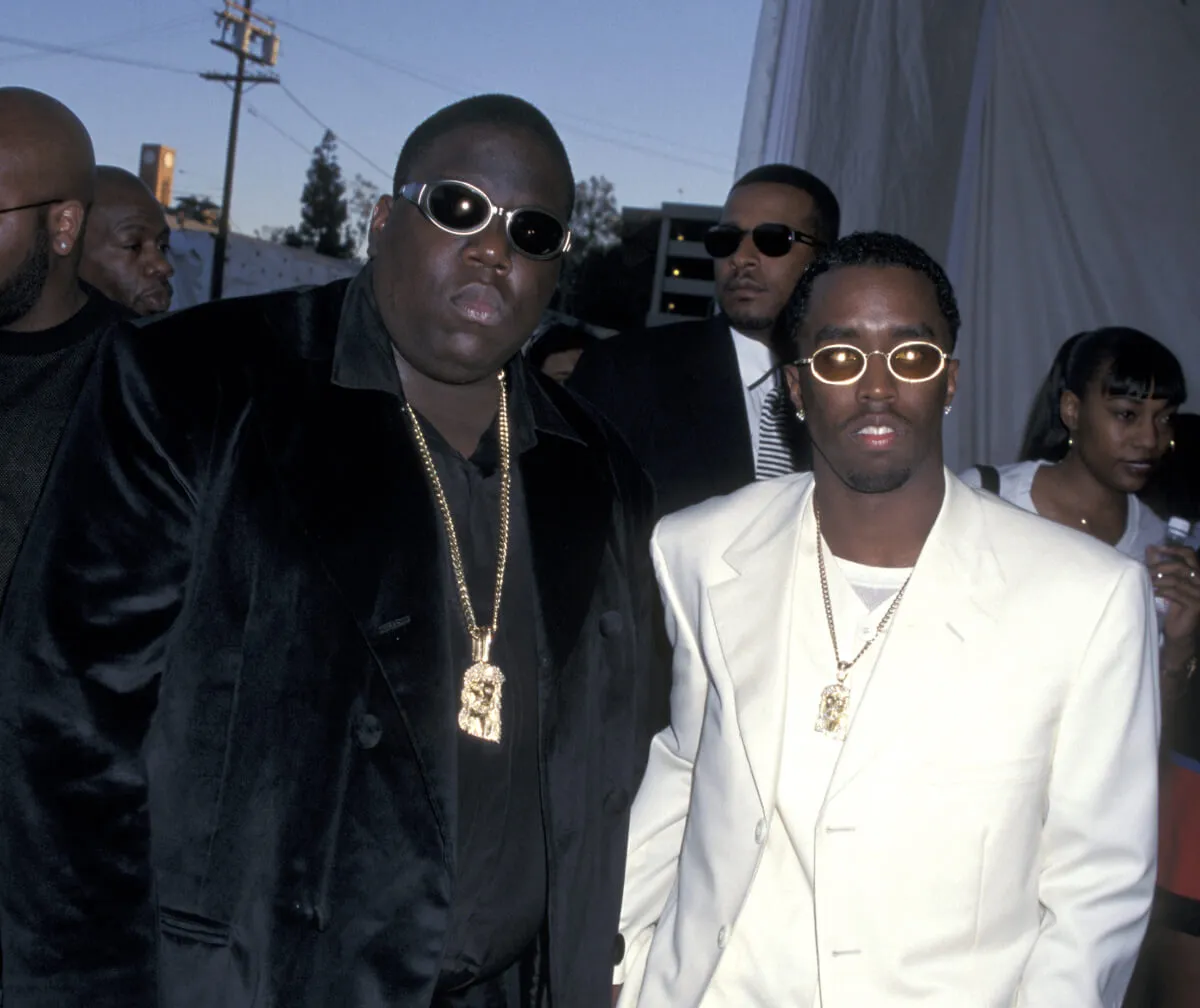 The Notorious B.I.G. wears a black suit and sunglasses and stands with Sean 'Diddy' Combs. Combs wears a white suit and sunglasses.