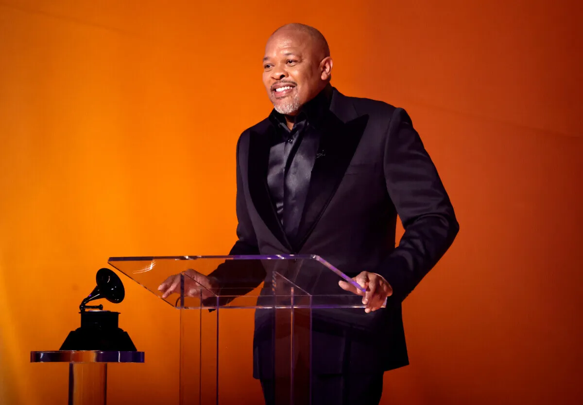 Dr. Dre wears a black suit and stands at a podium. A Grammy sits on a glass table next to him.