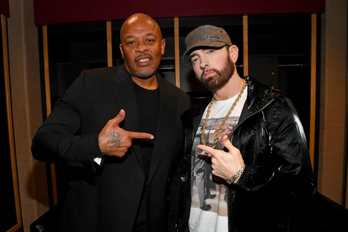 Dr. Dre wears black and stands with Eminem, who wears a black jacket and a white shirt. They point at each other.